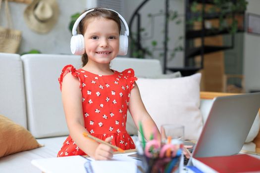 Smiling little girl in headphones handwrite study online using laptop at home, cute happy small child in earphones take Internet web lesson or class on PC