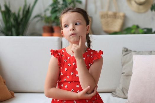 Pensive little girl sitting on sofa at living room. Daydreaming