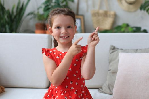 Cute little girl sitting on cozy sofa and pointing up on something intresting by fingers