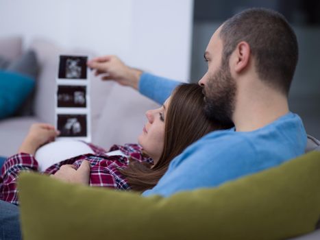 Young pregnant couple looking baby's ultrasound photo while relaxing on sofa at home