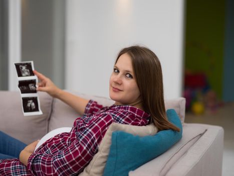 Young pregnant woman looking baby's ultrasound photo while relaxing on sofa at home