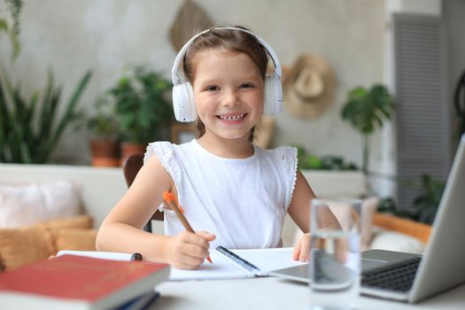 Smiling little girl in headphones handwrite study online using laptop at home, cute happy small child in earphones take Internet web lesson or class on PC