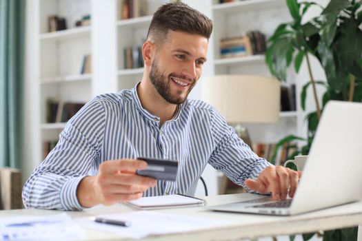 Smiling man sitting in office and pays by credit card with his laptop