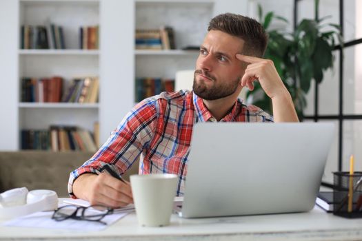 Unhappy frustrated young male holding head by hands sitting with laptop behind desk at home