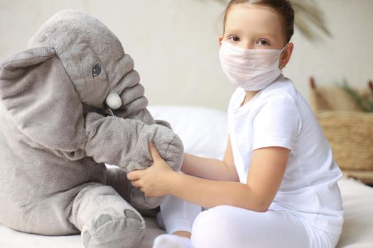 Stay at home quarantine coronavirus pandemic prevention. Sad child in protective medical mask and his elephant both in protective medical mask sit on bed