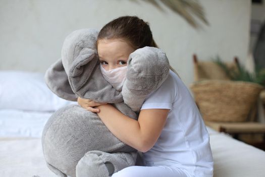 Stay at home quarantine coronavirus pandemic prevention. Sad child in protective medical mask and his elephant both in protective medical mask sit on bed