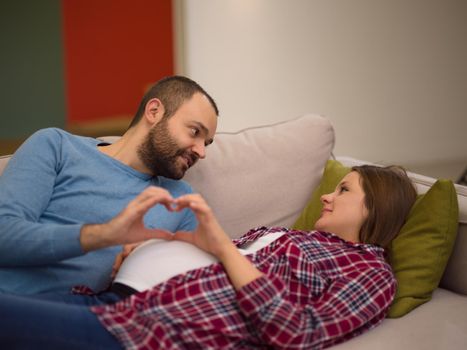 Happy man and pregnant woman showing heart sign with fingers while relaxing on the sofa at home