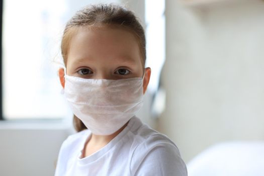 Little european girl wearing mask for protect Covid-19. Sick child Little girls look at the window. Copy space. sadness child at home in isolation