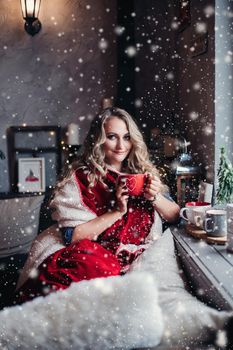 Attractive blonde lady wrapped in blankets felling snuggly while drinking hot tea by the window. Holiday concept