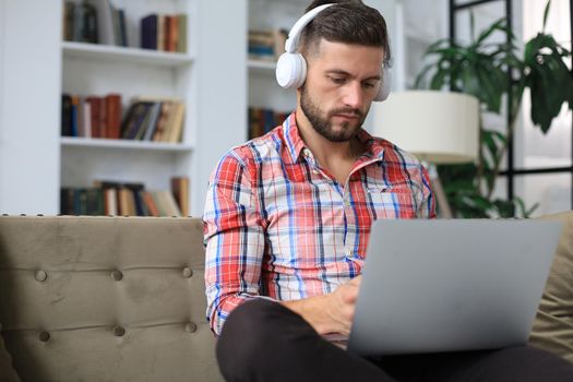 Concentrated young freelancer businessman sitting on sofa with laptop, working remotely online at home