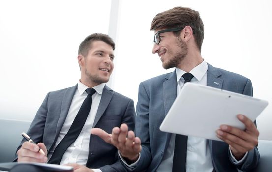 two young people are sitting in the office. One holds a tablet and gesticulates. The other holds a paper and a pen and listens.