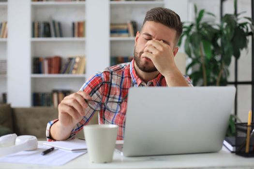 Unhappy frustrated young male holding head by hands sitting with laptop behind desk at home