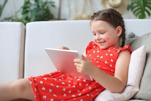 Smiling little girl lying on sofa playing online games, web surfing information, using funny applications on tablet