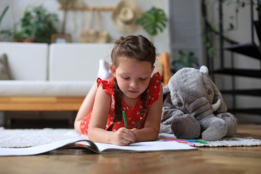 Smilling happy girl lying on warm floor with a toy elephant near to her enjoying creative activity, drawing pencils coloring pictures in albums