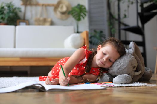 Smilling happy girl lying on warm floor with a toy elephant near to her enjoying creative activity, drawing pencils coloring pictures in albums