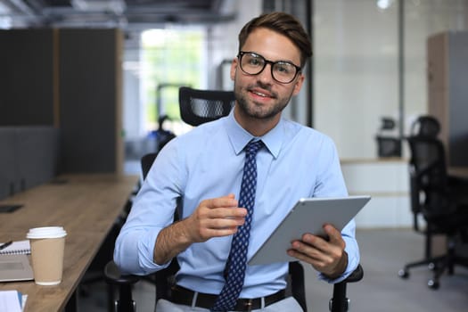 Handsome young businessman working using digital tablet and looking at camera while sitting in the office