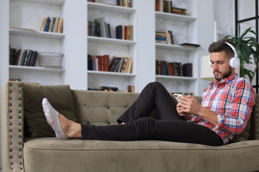 Attractive young man relaxing on a couch at home and using mobile phone for cheking social nets