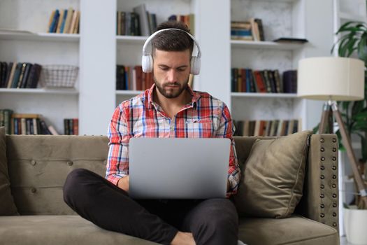 Concentrated young freelancer businessman sitting on sofa with laptop, working remotely online at home