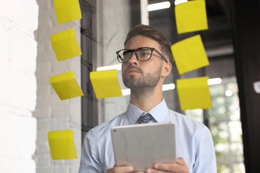 Young modern business man using adhesive notes while standing behind the glass wall in the office