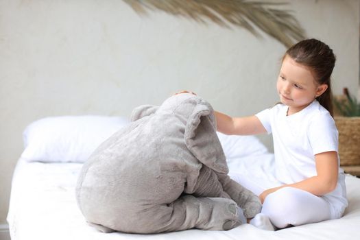 Sweet little girl sitting on her bed at home with toy elephant