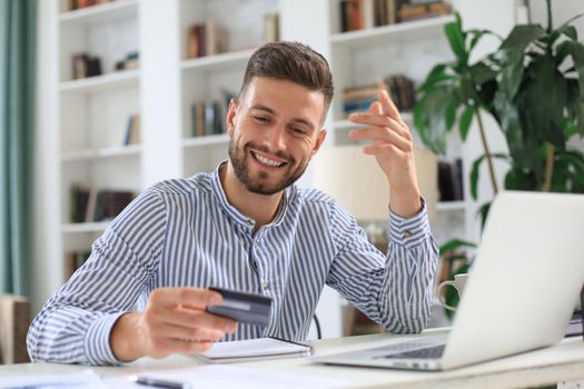 Smiling man sitting in office and pays by credit card with his laptop