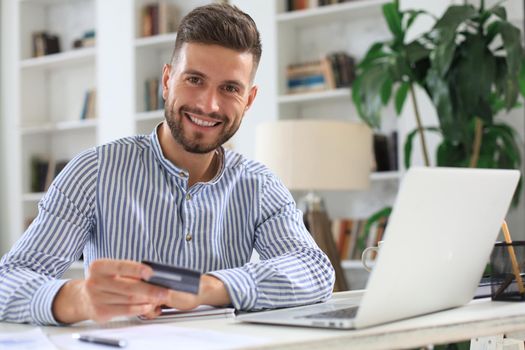 Smiling man sitting in office and pays by credit card with his laptop
