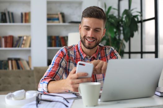 Attractive young man sitting at desk at home office and using mobile phone for cheking social nets