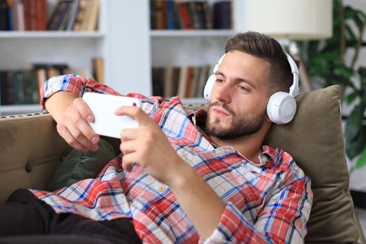 Attractive young man relaxing on a couch at home, listening music and playing on smartphone