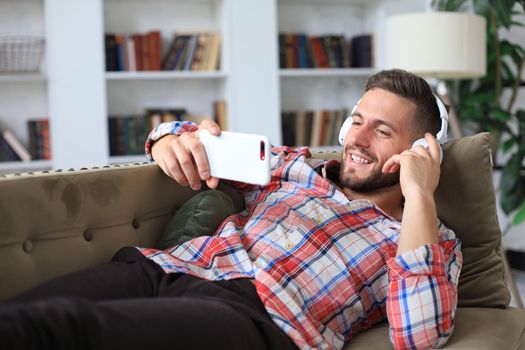 Attractive young man relaxing on a couch at home, listening music and playing on smartphone