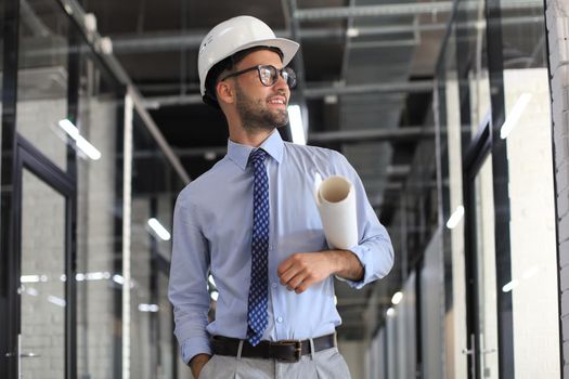 Confident young business man in hardhat holding blueprint and looking away while standing indoors