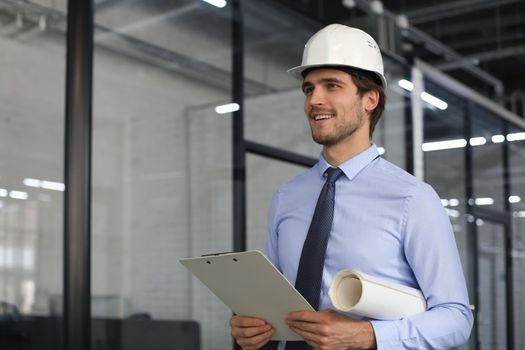 Confident young business man in hardhat holding blueprint and looking away while standing indoors