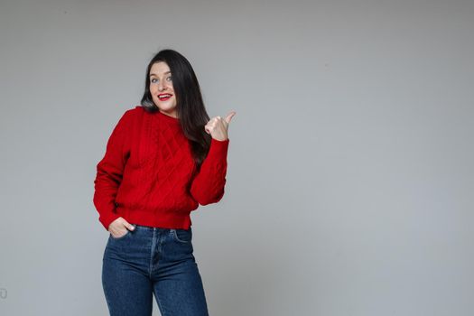 Stock photo of attractive brunette woman with long straight hair wearing red knitted sweater and jeans, pointing at blank space with thumb.