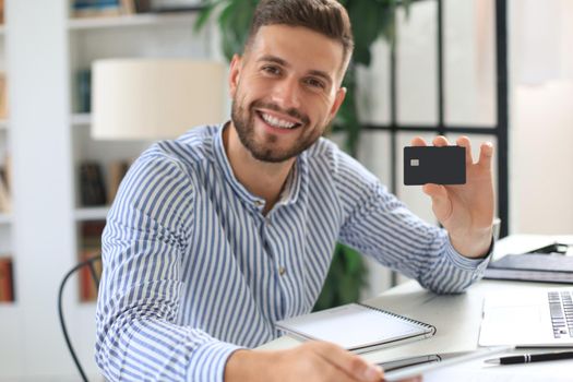 Smiling man sitting in office and pays by credit card with his laptop