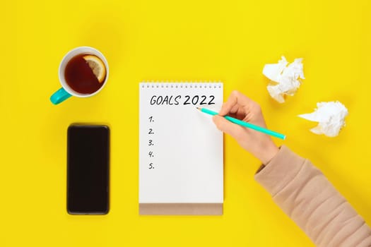 2022 goals banner with copy space. Notepad, pencil and cup of tea on a yellow background near crumpled sheets of paper and mobile phone. Copy space, Flat lay. Women hand write goals for 2020 year.