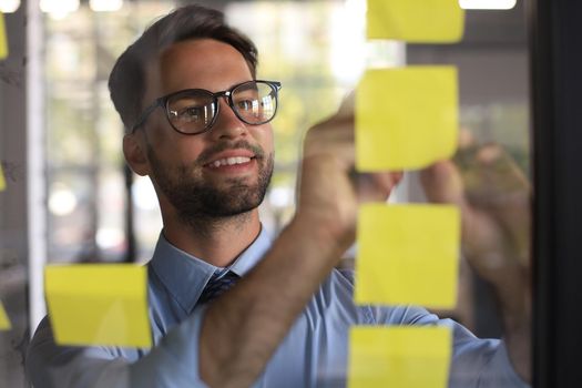 Young modern business man using adhesive notes while standing behind the glass wall in the office