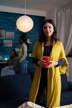 Portrait of smiling woman looking into camera holding plastic cup with beer celebrating friend birthday enjoying spending time together. Group of multi-ethnic people enjoying home party