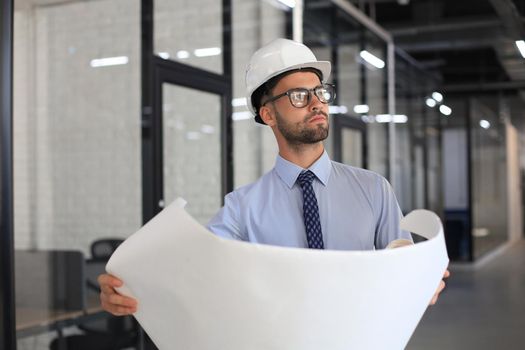 Confident young architect in hardhat holding a blueprint and looking at area for work