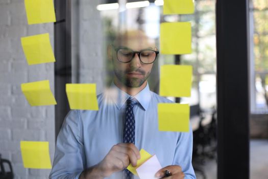 Young modern business man using adhesive notes while standing behind the glass wall in the office