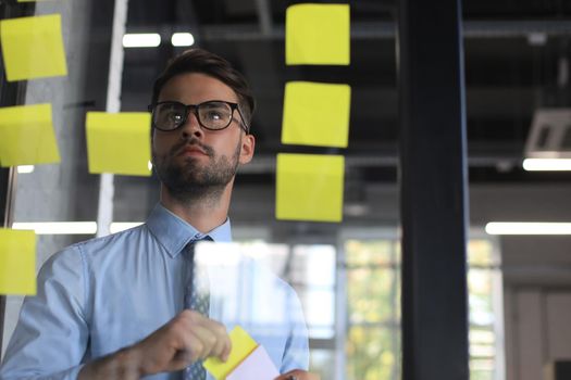 Young modern business man using adhesive notes while standing behind the glass wall in the office