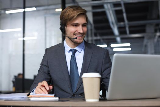 Smiling male call-center operator with headphones sitting at modern office, consulting online information in a laptop, looking up information in a file in order to be of assistance to the client