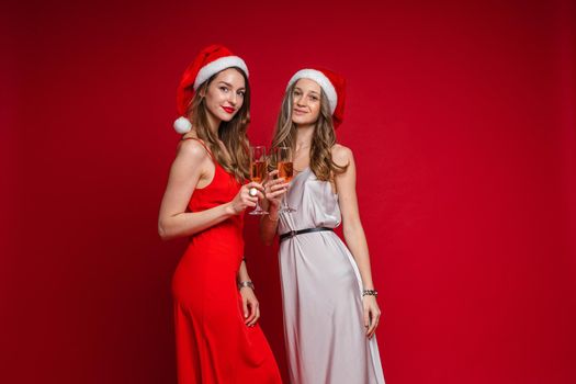 Studio portrait of two attractive girlfriends with long wavy hair in red and white silk dresses posing in Santa hats with glasses of rose wine. Isolated on red background.