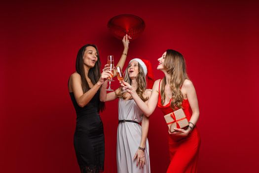 Full length of smiling pretty three girlfriends clinking glasses with champagne, isolated on red background. Party concept