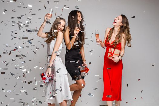 Portrait of jovial carefree beautiful ladies in cocktail dresses with glasses of alcoholic drinks dancing and having fun under falling silver confetti. Girls with drinks holding wrapped gifts.