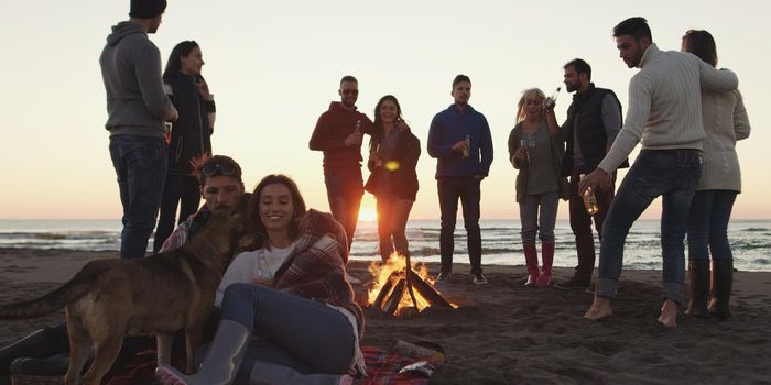 Happy Carefree Young Friends Having Fun And Drinking Beer By Bonefire On The Beach As The Sun Begins To Set