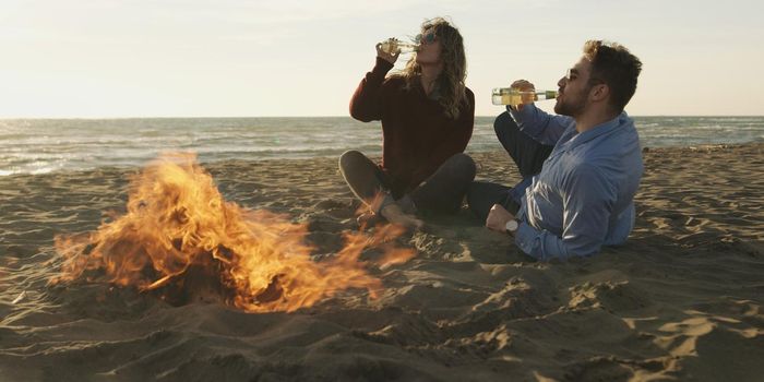 Young Couple Relaxing By The Fire, Drinking A Beer Or A Drink From The Bottle.