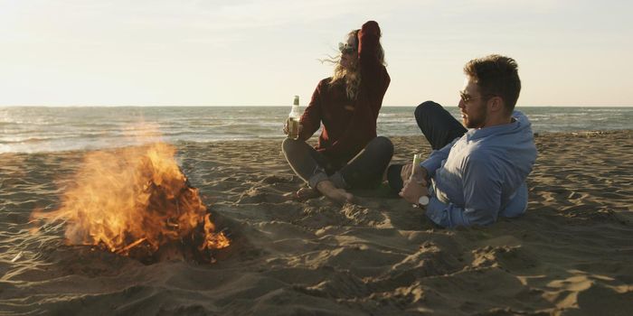 Young Couple Relaxing By The Fire, Drinking A Beer Or A Drink From The Bottle.