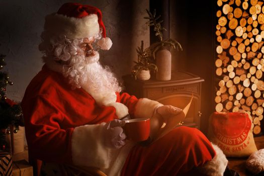Portrait of happy Santa Claus sitting at his room at home near fireplace and reading Christmas letter or wish list
