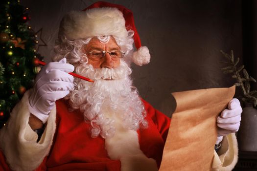 Portrait of happy Santa Claus sitting at his room at home near Christmas tree and reading Christmas letter or wish list