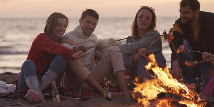 Group of young friends sitting by the fire late at night, grilling sausages and drinking beer, talking and having fun