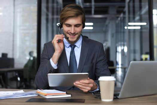 Smiling male business consultant with headphones sitting at modern office, video call looking at laptop screen. Man customer service support agent helpline talking online chat